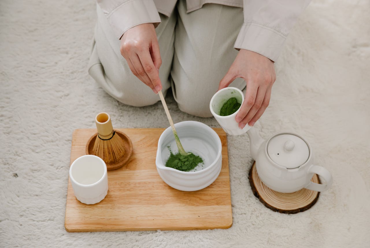 Hands carefully preparing matcha tea with traditional tools on a wooden tray.